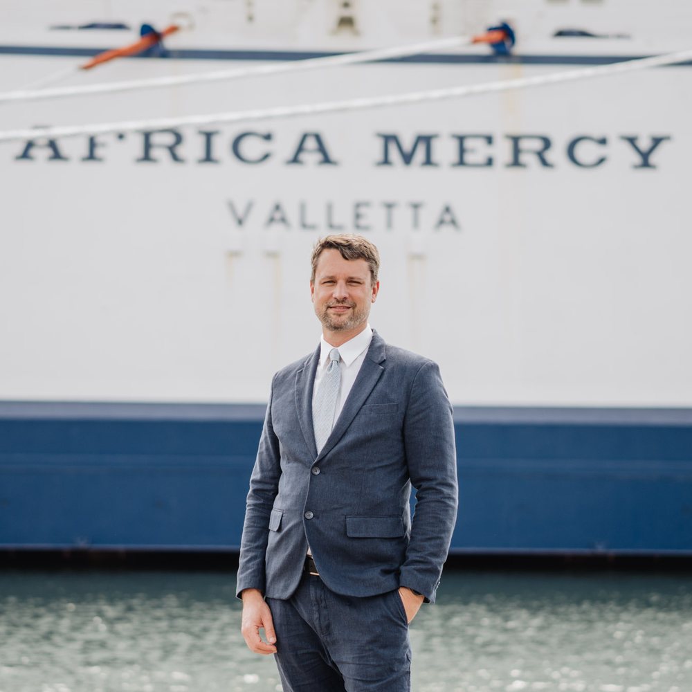 Dan Croutch in a navy suit, standing on the dock in front of the hospital ship Africa Mercy